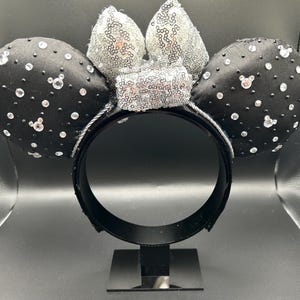 May include: Black satin mouse ears with clear rhinestones and a silver sequin bow. The ears are embellished with small, sparkling rhinestones. The headband is black and has a silver trim. A fun accessory for themed events.