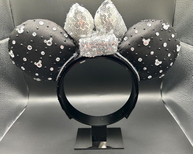 May include: Black satin mouse ears with clear rhinestones and a silver sequin bow. The ears are embellished with small, sparkling rhinestones. The headband is black and has a silver trim. A fun accessory for themed events.