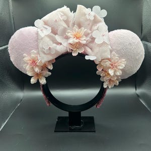 Op de afbeelding: Een haarband met twee grote, pluizige, lichtroze ronde oren. Een centrale, glinsterende, lichtroze strik is versierd met meerdere gelaagde witte en lichtroze stoffen bloemblaadjes. Extra trossen lichtroze bloesems sieren de onderkant.