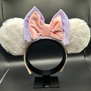 May include: A pair of mouse ear headbands with white fluffy ears, a lavender bow, and a pink velvet bow in the center. The headband is black with a sequined trim. The ears are round and fluffy.