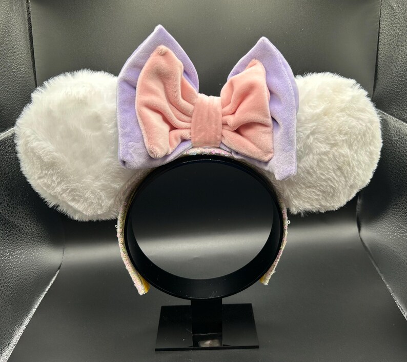 May include: A pair of mouse ear headbands with white fluffy ears, a lavender bow, and a pink velvet bow in the center. The headband is black with a sequined trim. The ears are round and fluffy.