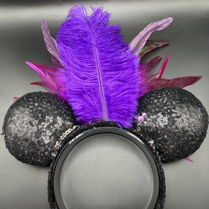 May include: A headband featuring two round, black sequined ears and a central arrangement of vibrant purple and magenta feathers. A small, light purple, circular accent is attached to the base of the right ear, adding a subtle detail to the design.