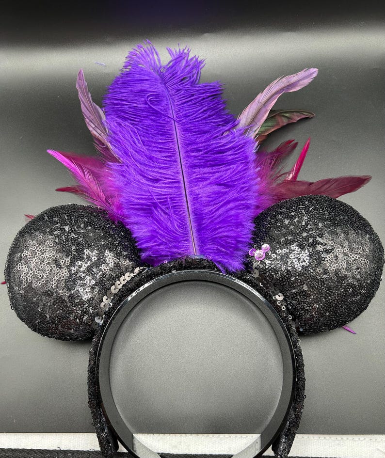May include: A headband featuring two round, black sequined ears and a central arrangement of vibrant purple and magenta feathers. A small, light purple, circular accent is attached to the base of the right ear, adding a subtle detail to the design.