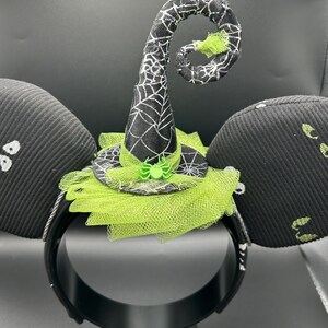 May include: Black Halloween-themed headband with witch hat and mouse ears. The witch hat is black with a white spiderweb design and a green spider. The ears are black with white ghost designs.