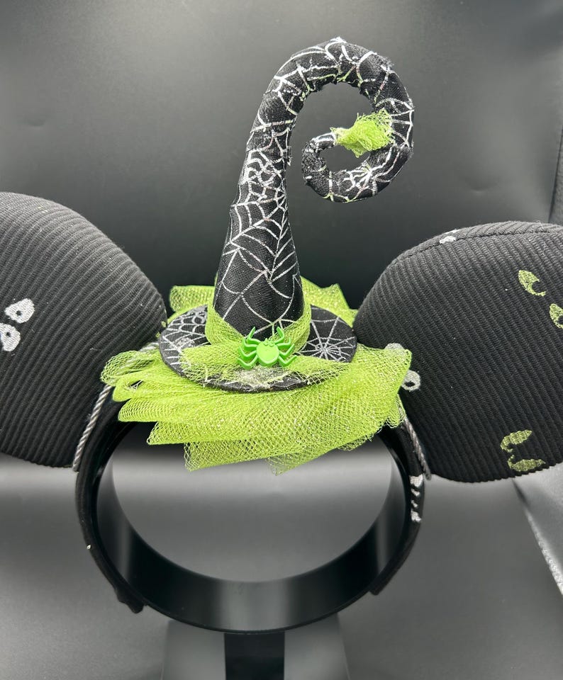 May include: Black Halloween-themed headband with witch hat and mouse ears. The witch hat is black with a white spiderweb design and a green spider. The ears are black with white ghost designs.