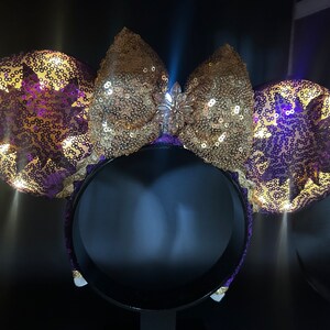 Pu&ograve; includere: Orecchie di Minnie Mouse in paillettes viola e dorate con un fiocco dorato e un gioiello scintillante al centro. Le orecchie hanno la forma del sole.