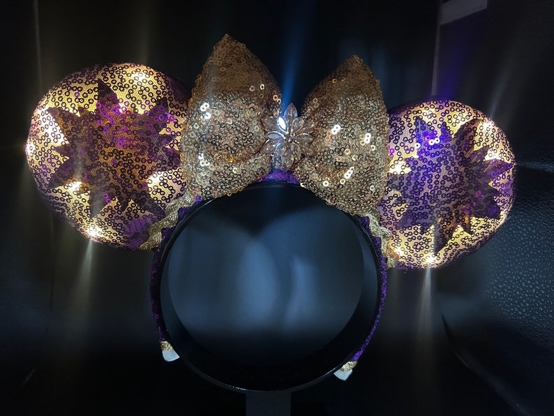 Pu&ograve; includere: Orecchie di Minnie Mouse in paillettes viola e dorate con un fiocco dorato e un gioiello scintillante al centro. Le orecchie hanno la forma del sole.