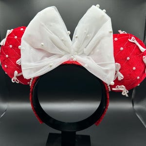 May include: Red velvet mouse ear headband with a large white bow adorned with faux pearls. The ears are embellished with white bows and pearl accents. The headband is black.