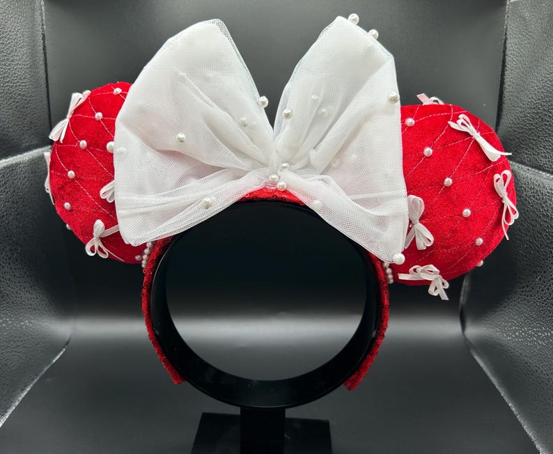 May include: Red velvet mouse ear headband with a large white bow adorned with faux pearls. The ears are embellished with white bows and pearl accents. The headband is black.