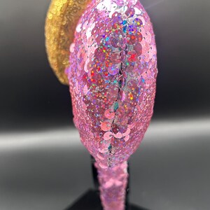 May include: A headband featuring two rounded ears covered in shimmering sequins. One ear is a bright metallic gold, while the other is a vibrant pink with iridescent flecks. The headband base is also fully covered in matching pink sequins.