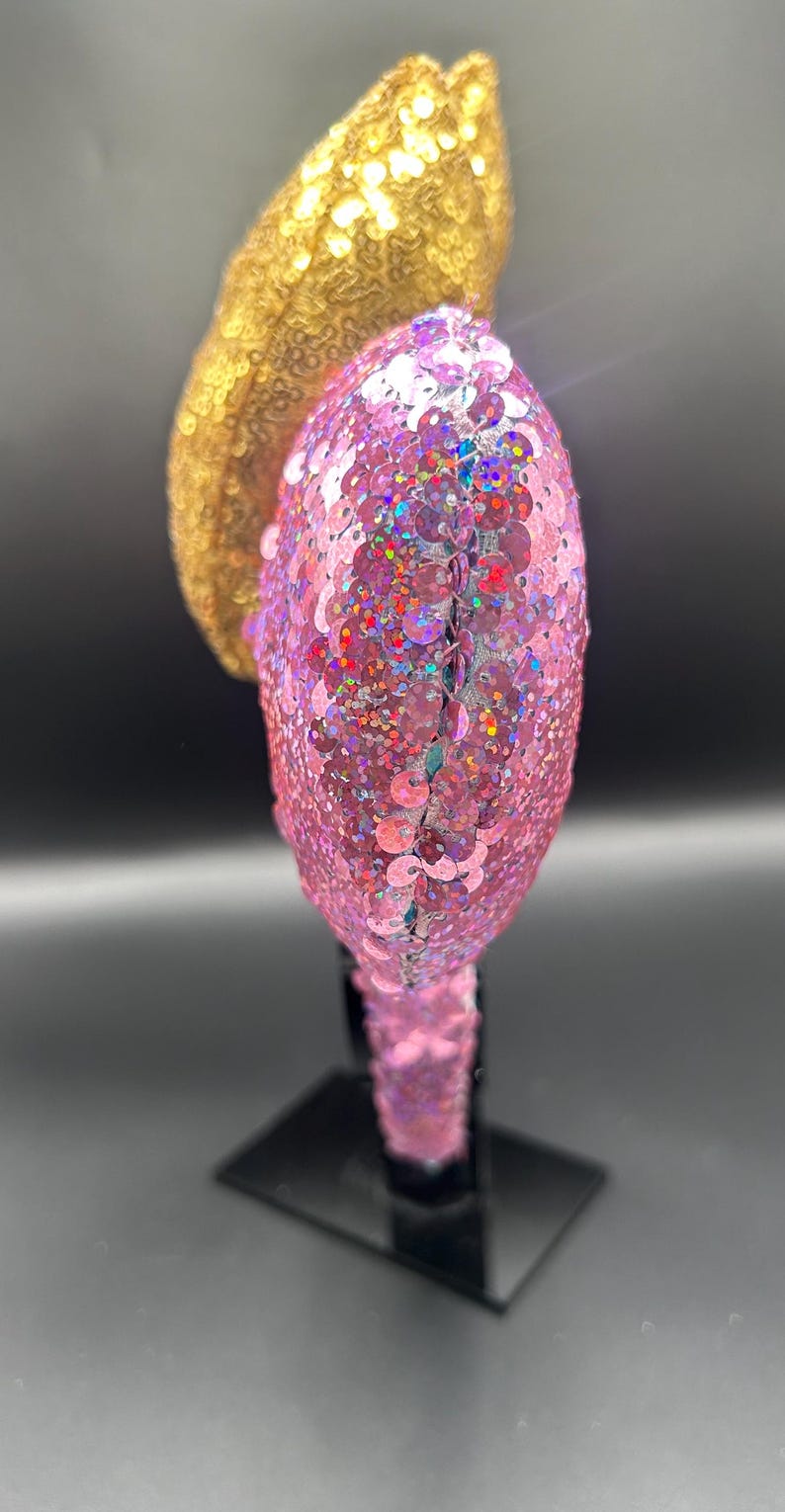 May include: A headband featuring two rounded ears covered in shimmering sequins. One ear is a bright metallic gold, while the other is a vibrant pink with iridescent flecks. The headband base is also fully covered in matching pink sequins.