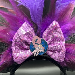 May include: A headband featuring black sequined ears, a large light purple sequined bow, and vibrant purple and magenta feathers. A circular enamel pin depicting a cartoon character with pale skin, long pink hair, and a wide grin is centered on the bow.