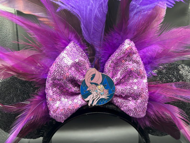 May include: A headband featuring black sequined ears, a large light purple sequined bow, and vibrant purple and magenta feathers. A circular enamel pin depicting a cartoon character with pale skin, long pink hair, and a wide grin is centered on the bow.