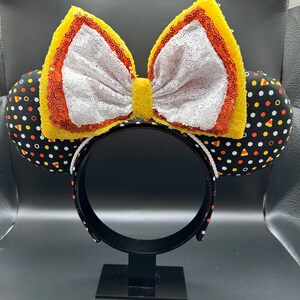 May include: A Halloween-themed headband with black ears patterned with white polka dots and candy corn. The large bow is white with yellow and orange sequin trim. The headband is black with a white rope trim.