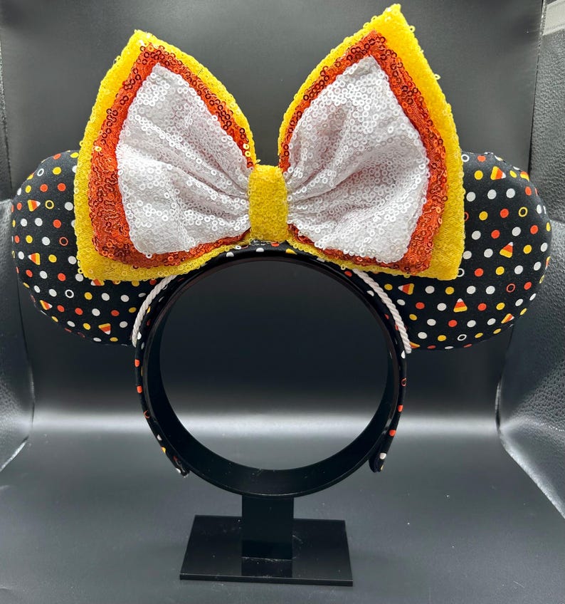 May include: A Halloween-themed headband with black ears patterned with white polka dots and candy corn. The large bow is white with yellow and orange sequin trim. The headband is black with a white rope trim.