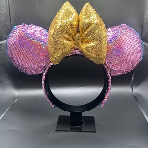 May include: A headband featuring two circular ears covered in iridescent pink sequins and a large, central bow covered in shimmering gold sequins. The headband frame is black, and the inner band is lined with matching pink sequins for a cohesive look.