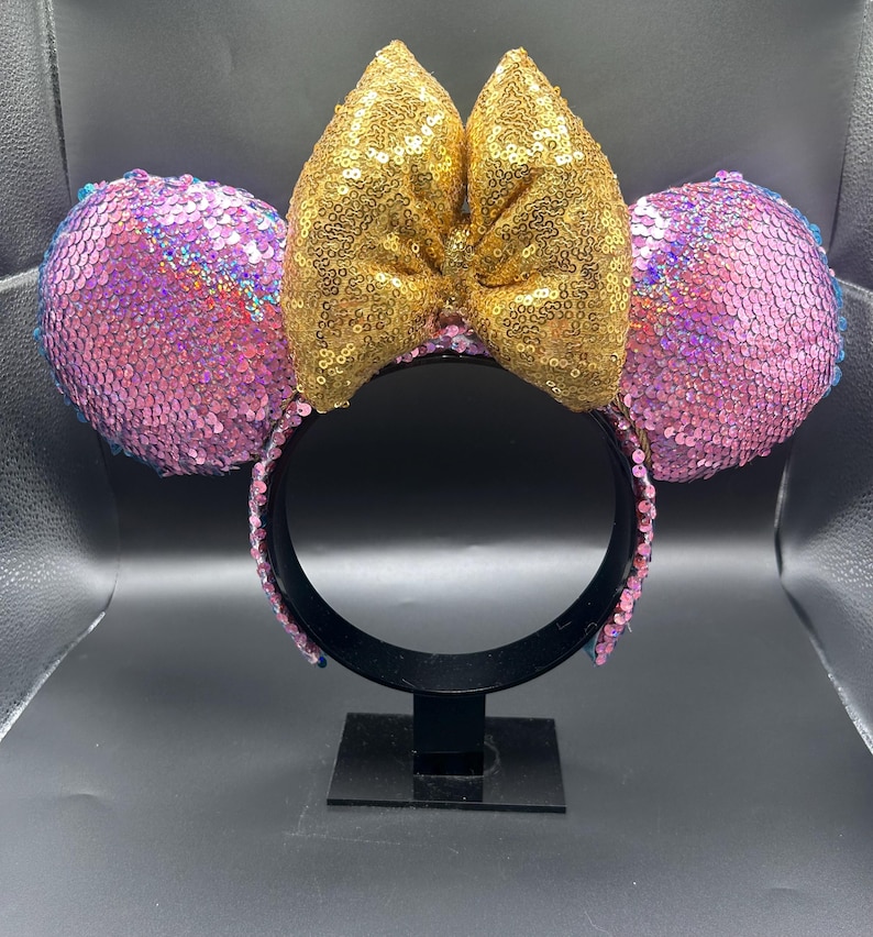 May include: A headband featuring two circular ears covered in iridescent pink sequins and a large, central bow covered in shimmering gold sequins. The headband frame is black, and the inner band is lined with matching pink sequins for a cohesive look.