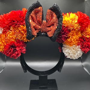 May include: A headband featuring two large circular clusters of artificial flowers in shades of red, orange, yellow, and white. Centered between the floral clusters is a large bow made of shimmering orange sequins with black sequined edges.