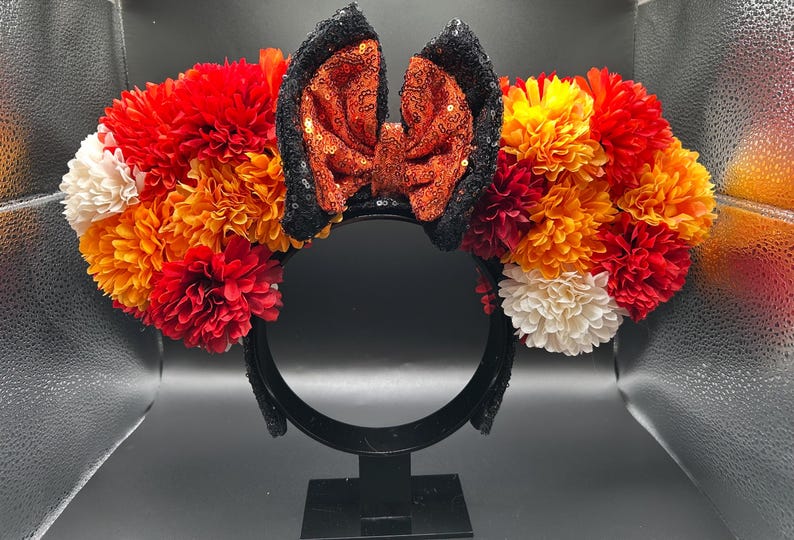 May include: A headband featuring two large circular clusters of artificial flowers in shades of red, orange, yellow, and white. Centered between the floral clusters is a large bow made of shimmering orange sequins with black sequined edges.