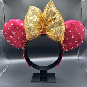 May include: A red headband with two round, red ears adorned with clear and gold-colored embellishments. A large, gold sequined bow sits between the ears. The headband is displayed on a black stand.