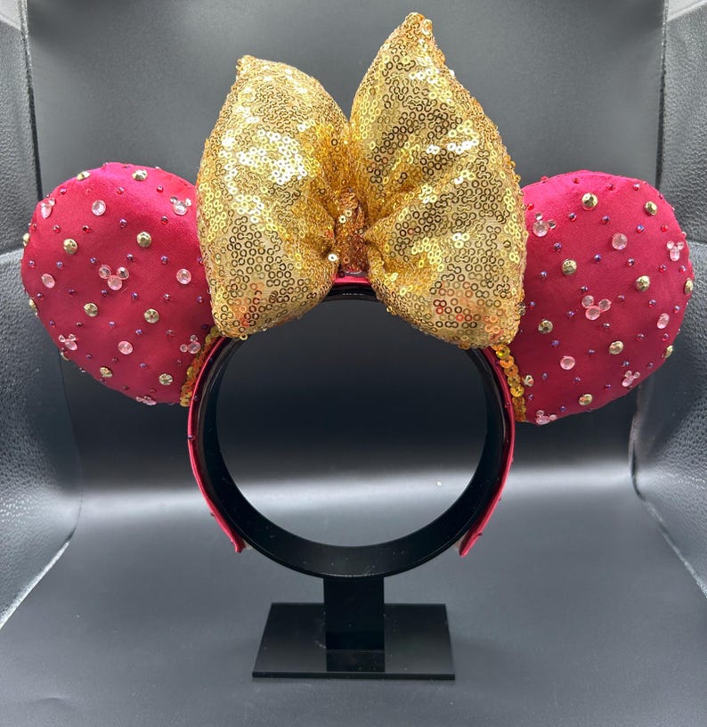 May include: A red headband with two round, red ears adorned with clear and gold-colored embellishments. A large, gold sequined bow sits between the ears. The headband is displayed on a black stand.