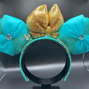 May include: A teal sequined headband with two teal ears and a gold sequined bow. Each ear has a teal bow with a rhinestone centre. The headband is trimmed with gold rope and has a black interior.