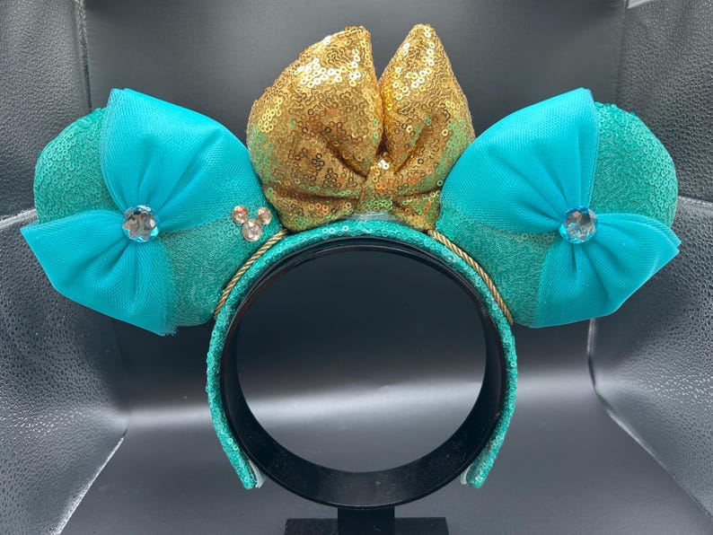 May include: A teal sequined headband with two teal ears and a gold sequined bow. Each ear has a teal bow with a rhinestone centre. The headband is trimmed with gold rope and has a black interior.