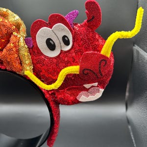 May include: A red sequined dragon headband with a yellow pipe cleaner mustache and eyebrows. The dragon has large white eyes with black pupils, and pink glitter accents. The headband is black.