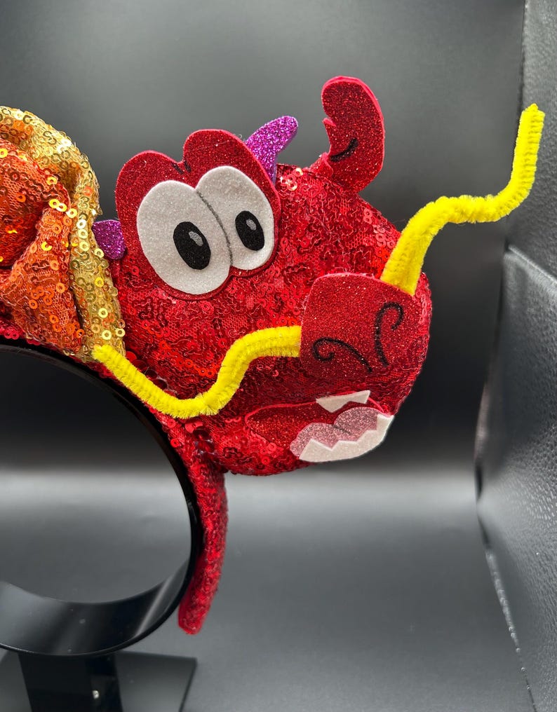 May include: A red sequined dragon headband with a yellow pipe cleaner mustache and eyebrows. The dragon has large white eyes with black pupils, and pink glitter accents. The headband is black.