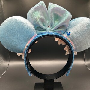 Peut inclure: Un serre-t&ecirc;te avec deux grandes oreilles circulaires recouvertes de velours bleu clair et un n&oelig;ud central bleu clair iris&eacute;. L'arceau est recouvert de sequins bleu fonc&eacute;, agr&eacute;ment&eacute; de petites fleurs blanches et d'une minuscule ic&ocirc;ne bleue.
