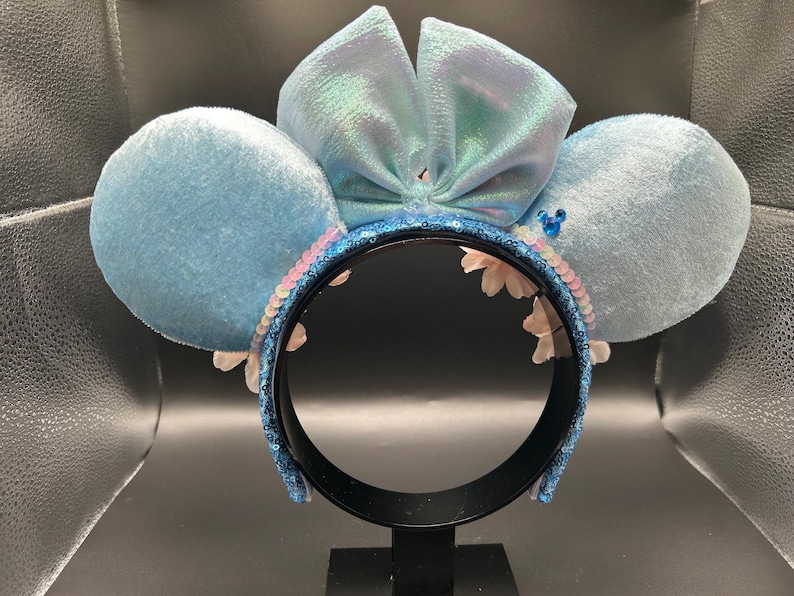 Peut inclure: Un serre-t&ecirc;te avec deux grandes oreilles circulaires recouvertes de velours bleu clair et un n&oelig;ud central bleu clair iris&eacute;. L'arceau est recouvert de sequins bleu fonc&eacute;, agr&eacute;ment&eacute; de petites fleurs blanches et d'une minuscule ic&ocirc;ne bleue.