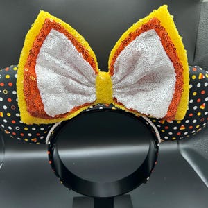 May include: A festive headband featuring mouse ears and a large bow. The ears are black with a pattern of candy corn, while the bow is yellow, orange, and white with sequins. The headband itself is black.