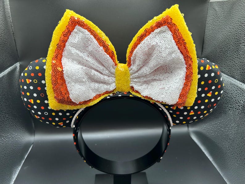 May include: A festive headband featuring mouse ears and a large bow. The ears are black with a pattern of candy corn, while the bow is yellow, orange, and white with sequins. The headband itself is black.