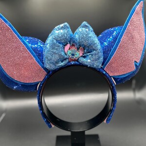 May include: A blue sequined headband with large, pink glitter-covered ears and a blue sequined bow. The bow features a small character figure. The headband is designed to resemble a popular animated character.