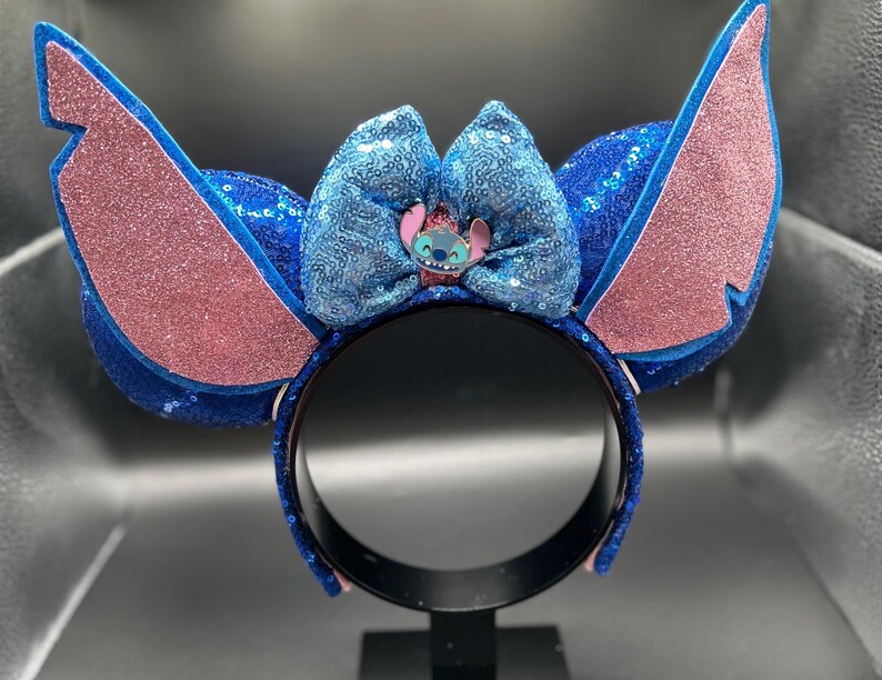 May include: A blue sequined headband with large, pink glitter-covered ears and a blue sequined bow. The bow features a small character figure. The headband is designed to resemble a popular animated character.