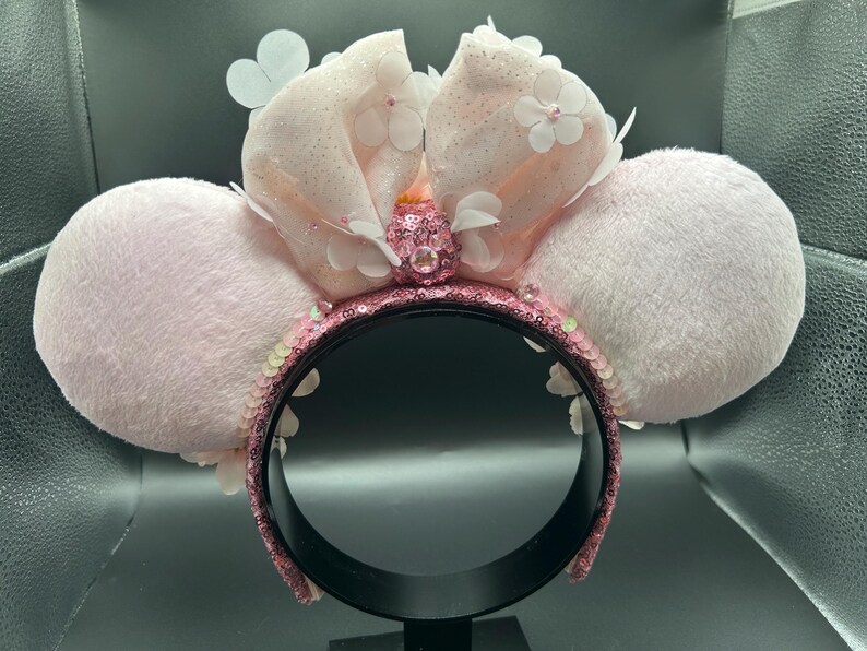 Op de afbeelding: Een haarband met twee grote, pluche, lichtroze ronde oren. Een centrale, glinsterende, lichtroze stoffen strik is versierd met kleine witte bloemapplicaties en een centrale roze edelsteen. Het frame is bedekt met glinsterende roze lovertjes.