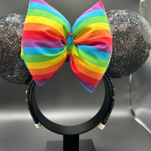 K&ouml;nnte beinhalten: Schwarze Pailletten-Minnie-Maus-Ohren mit einer gro&szlig;en regenbogenfarbenen Schleife.