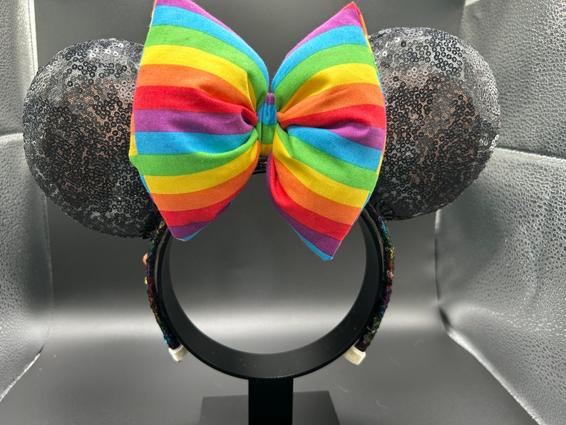 K&ouml;nnte beinhalten: Schwarze Pailletten-Minnie-Maus-Ohren mit einer gro&szlig;en regenbogenfarbenen Schleife.