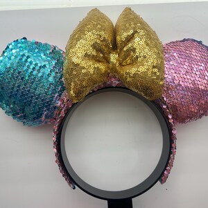 May include: A headband featuring two circular ears covered in sequins, with a bright blue ear on the left and a pink ear on the right. A large, central bow made of gold sequins sits between the ears. The headband frame is black and circular.