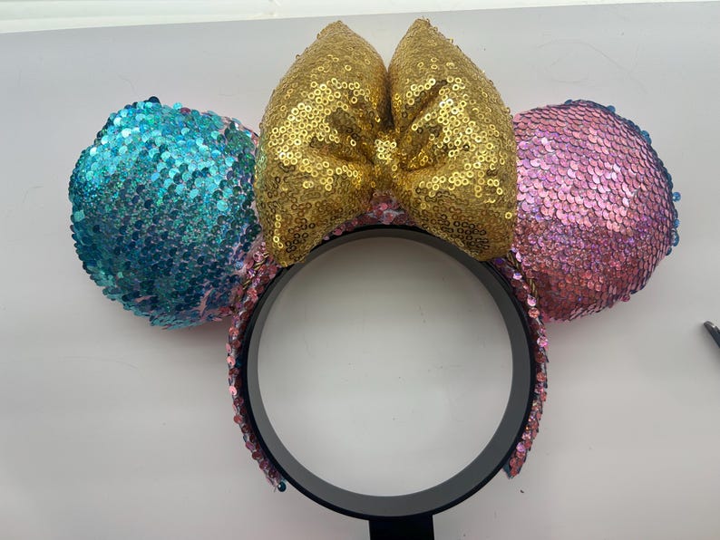 May include: A headband featuring two circular ears covered in sequins, with a bright blue ear on the left and a pink ear on the right. A large, central bow made of gold sequins sits between the ears. The headband frame is black and circular.