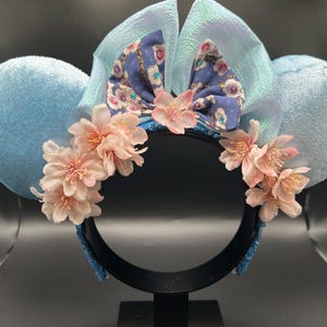 Peut inclure: Un serre-t&ecirc;te avec deux grandes oreilles en velours bleu clair, un n&oelig;ud central bleu iris&eacute; et une composition d&eacute;corative de fleurs de cerisier artificielles rose p&acirc;le fix&eacute;es sur le devant de la monture noire.