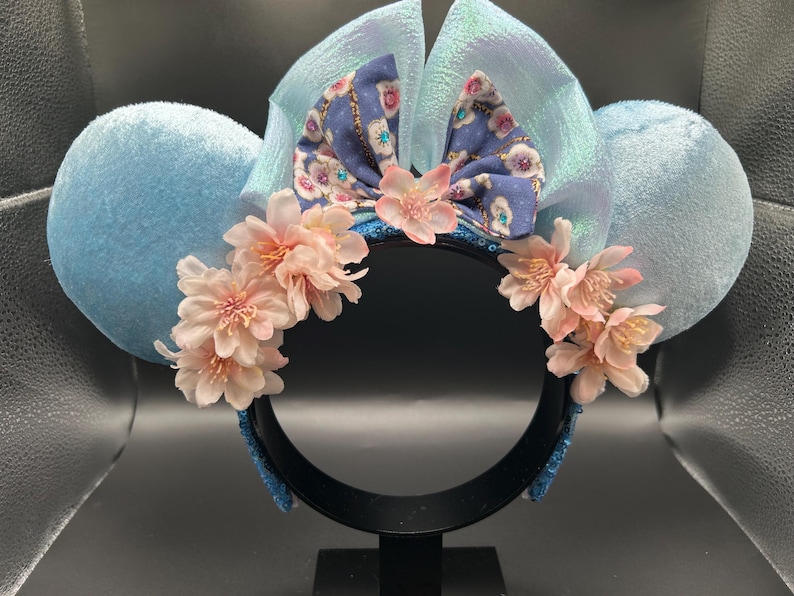 Peut inclure: Un serre-t&ecirc;te avec deux grandes oreilles en velours bleu clair, un n&oelig;ud central bleu iris&eacute; et une composition d&eacute;corative de fleurs de cerisier artificielles rose p&acirc;le fix&eacute;es sur le devant de la monture noire.