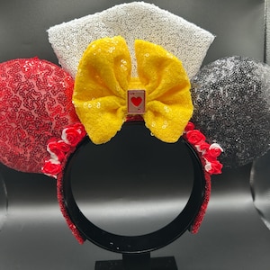 May include: A headband featuring two circular ears covered in red and black sequins, a central yellow fabric bow with a small playing card charm depicting a red heart, a white sequined fan-shaped accent, and small red and white fabric roses.