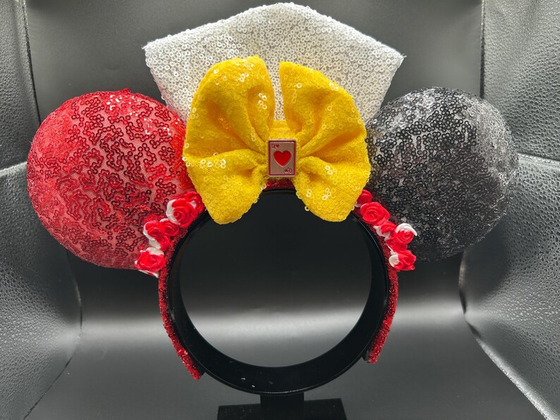 May include: A headband featuring two circular ears covered in red and black sequins, a central yellow fabric bow with a small playing card charm depicting a red heart, a white sequined fan-shaped accent, and small red and white fabric roses.