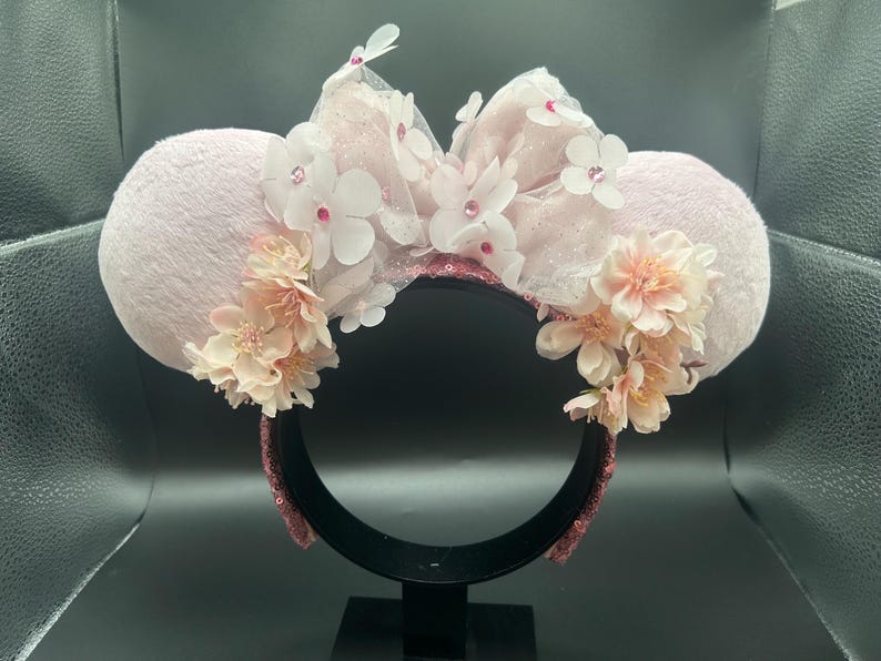 May include: A light pink Minnie Mouse ear headband. The ears are decorated with a large bow made of sheer fabric, white flowers, and pink sequins. Clusters of peach-coloured flowers are also attached to the ears.