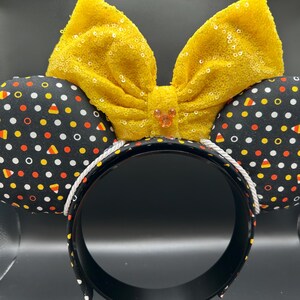 May include: A festive headband featuring black ears patterned with white polka dots, orange, yellow, and candy corn shapes. A large, yellow sequined bow sits atop the headband, with a small jewel in the center. The headband is black with a white rope trim.
