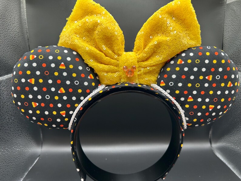 May include: A festive headband featuring black ears patterned with white polka dots, orange, yellow, and candy corn shapes. A large, yellow sequined bow sits atop the headband, with a small jewel in the center. The headband is black with a white rope trim.