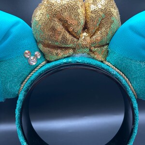 May include: Teal mouse ear headband with a gold sequin bow and teal bows. The headband has a gold rope trim and sparkling jewel accents. A fun accessory for themed events.