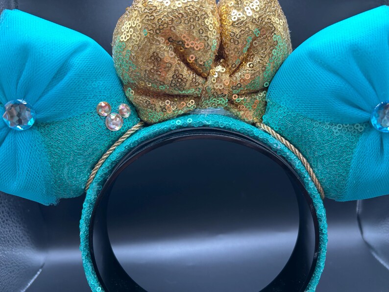 May include: Teal mouse ear headband with a gold sequin bow and teal bows. The headband has a gold rope trim and sparkling jewel accents. A fun accessory for themed events.