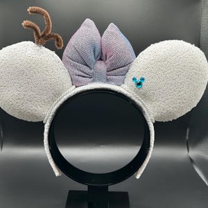 May include: A festive headband featuring large, sequined gray mouse ears. A shimmering bow in shades of purple and blue sits between the ears. Brown pipe cleaner antlers and a blue jewel accent complete the design. The headband is white.